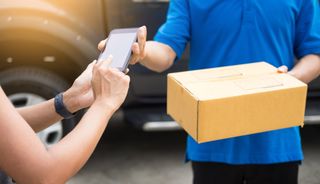 Woman tapping smartphone while delivery person hands her a package.