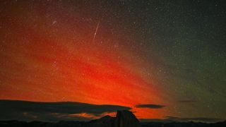 A Perseid meteor shoots through the sky as a red aurora dances near the horizon