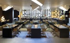 A restaurant with man long dark wooden tables, chairs, large roof down lighters and a large window.