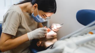 Dentist wearing a blue surgical mask and gloves leans over a patient's head to inspect his teeth with a mirror