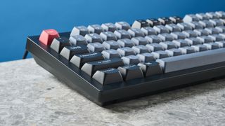 A photograph of the Keychron V6 in black, with gray , black and orange keycaps. The keyboard is positioned on a stone slate, with a blue wall in the background.