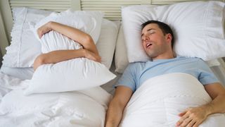 A man sleeping on his back snoring next to his partner who's covered her head and ears with a pillow