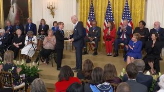 a man in a blue suit shakes hands with a woman in a dark blue suit in a crowded room with multiple american flags