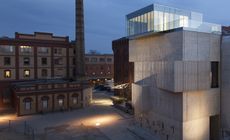 On the right of the image is the exterior of an abstractly designed sandy-coloured building with floor to ceiling glass design on the topmost layer. On the left is two-toned building in 2 shades of brown 