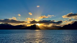 a bright orange sun peaks from behind the tip of a mountain and some clouds. Water below and blue sky above.