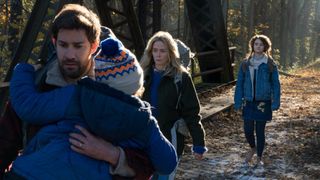A screenshot from a quiet place which shows a concerned-looking man carrying a baby with his family close behind