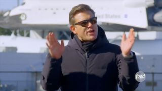 a man wearing sunglasses and a black jacket speaks and gesticulates, with a life-sized space shuttle orbiter model in the background