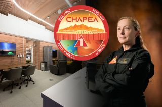 a woman in a black jumpsuit stands with arms crossed in front of a room with rolling chairs and conference tables