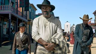 Idris Elba stands in a cowboy hat as Rufus Buck in The Harder They Fall with Regina King and LaKeith Stanfield on each side of him.