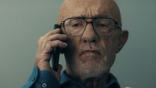 Close up of a bald, older man with a white goatee and wearing glasses. He is holding a cell phone up to his ear. He looks upset.