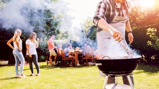 A man cooking on a grill with a people eating around a table in the background