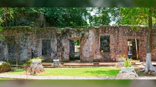 Casa de Hernan Cortes in Veracruz, Mexico. There are several N-shaped white stone structures covered in green moss. In the front there is a patch of green grass . In the background you can see lots of green trees.