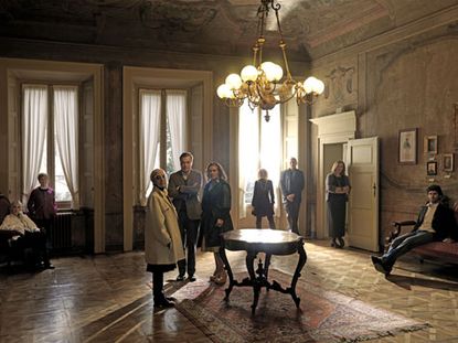 People standing in a room with a round table on a rug in the center next to large windows with shutters.