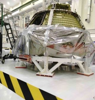 cone-shaped spacecraft on a white circular mount. the bottom is wrapped in plastic. a large facility with ladders and other equipment is in background. caution tape is in the front