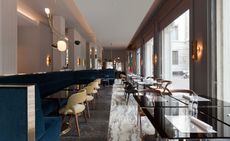 View of the interior of the restaurant with deep velvet blue and beige sitting area, gray and white marble flooring, and high windows.