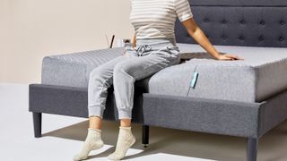 A woman wearing gray sweat pants sits on the edge of the Siena Memory Foam Mattress