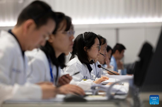 Six people in white outfits sitting in a mission control room, all facing the same direction.