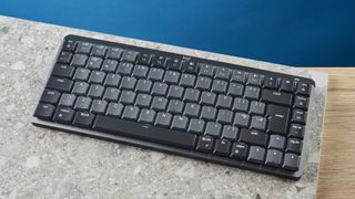 A photo of the Logitech MX Mechanical Mini keyboard in black and gray, on a stone slab and wooden table with a blue background.