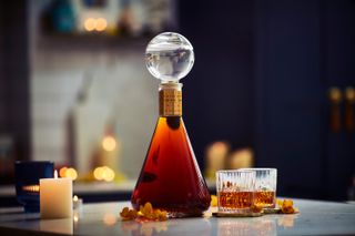 A bottle of Maestro Dobel 50 tequila on a light coloured reflective surface with candles, two filled glasses and small flowers