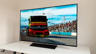 The LG OLED48C3 TV on a white table in front of a white wall. The screen shows a racing truck in front of a crowd on bleachers.