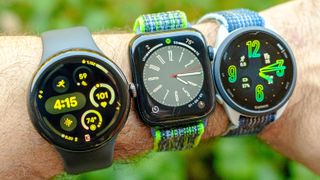 The Google Pixel Watch 3 in 45mm next to the Apple Watch SE 2022 and Garmin Forerunner 165 on a user's wrist with a green out of focus background