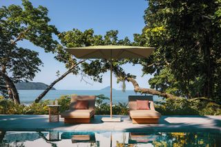 A square brown parasol with two loungers under it in front of a pool with a view of the ocean.