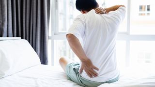 A man sits on the edge of the bed holding his neck and back