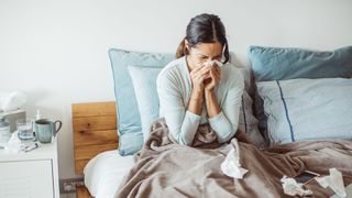 woman sitting up in bed blowing her nose. she's surrounded by used tissues and a cup of tea can be seen on a bedside table beside her