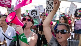 Picture of a crowd of people holding placards. Two activists are shown in the foreground of the image. On the left, an activist is shouting into a pink megaphone. The activist on the right is holding up a placard and shouting 