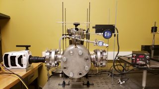 a table-sized metal chamber sealed with four large bolts, connected to other mechanical devices with tubes and wires across three different table surfaces in a room with yellow walls.