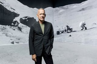 a man in a navy-blue jacket stands in front of a wall bearing imagery of the moon from NASA's Apollo program.