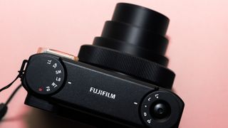 A photograph of the Fujifilm Instax mini 99 in black, set against a pink background.