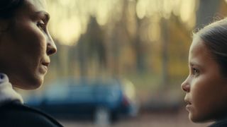 Close up of mother and young daughter looking at each other. The background is blurry, but the scene shows a big car and some woods.