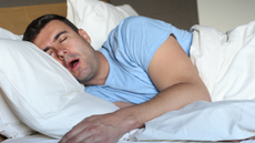 A man in a blue t shirt lying on his side in bed, sleeping with his mouth wide open