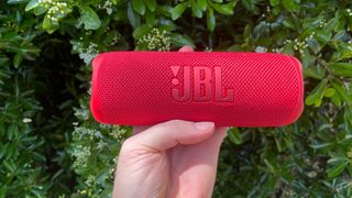 A red JBL Flip 6 speaker being held in front of a bush.