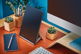 An orange desk with blue notepad and pencil placed on the pad. Green plants in 3 small wooden style pots. An ipad on an ipad stand with a peek of a white monitor on a monitor stand and white keyboard