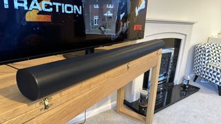 A black Sonos Arc soundbar on a wooden TV cabinet, in front of a TV showing Extraction 2 on Netflix. In the background is a fireplace and an armchair with a geometric pattern.