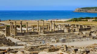 a large ruin containing numerous columns is in the foreground of the image with a beach in the background lined with parasols and people.