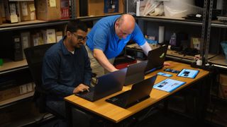 One person leans over a desk, another sits beside, while they both look at a desk with several laptops on top.