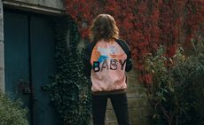 A female model standing in a garden, back turned to the camera, wearing a a top with block sleeves and a colourful body with the word "Baby" written across the back in a stencil font.