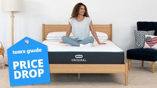 A woman with long curly dark hair smiles and sits cross-legged on top of a Bear Original memory foam mattress placed on a light wooden bedframe; a blue price drop sales badge is overlaid on the bottom left hand corner