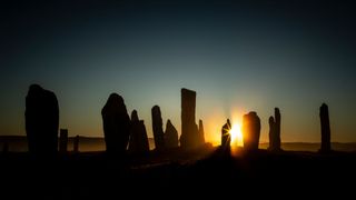 block shaped stones are visible against the sky in shadow. Between two of the stones, towards the right of the image, the sun rises. the rays of the sun shine against one of the stones.