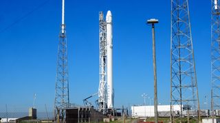 A skinny white rocket stands surrounded by metal-framed lighting towers. A tall streetlamp stands in the foreground. The sky is very blue.