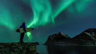 A surfer carrying a surfboard gazes at the aurora light show in the sky