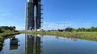 a silver, white and red rocket stands inside a large hangar behind a pond