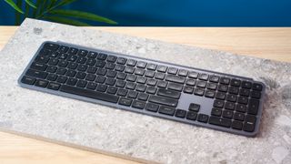 a black and grey budget wireless Bluetooth keyboard with a full-size layout and a numerical keypad is photographed against a blue background