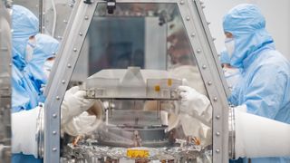 four people in blue protective equipment handle a metal canister inside a laboratory