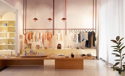 Lagalá shop IMOX, clothing hanging on copper rails suspended from ceiling with copper lamps above, wooden table and pot plant