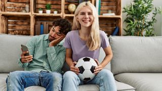 A woman smiles watching a soccer game, while a man next her sits looking bored on his phone