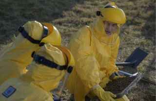 Three people in yellow hazmat suits find a stone tablet